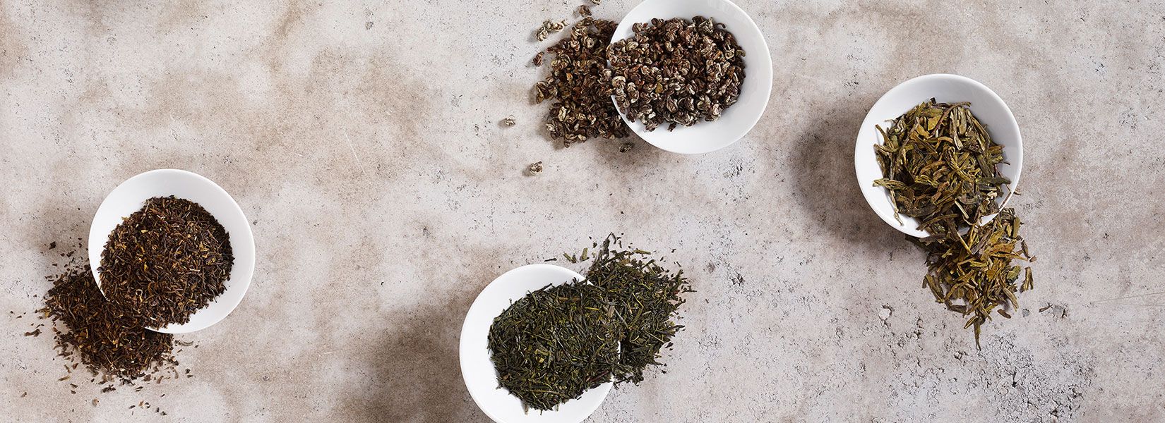 Various loose tea leaves in white bowls on a stone surface.