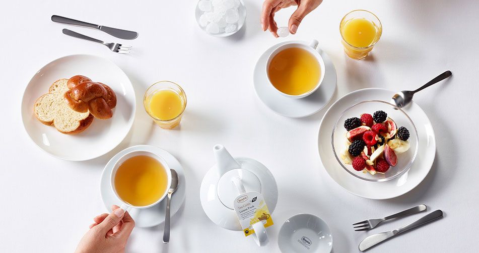 Breakfast table with cups of Ronnefeldt tea, braided bread, fresh fruit with yogurt, glasses of orange juice, and a white teapot on a white tablecloth.