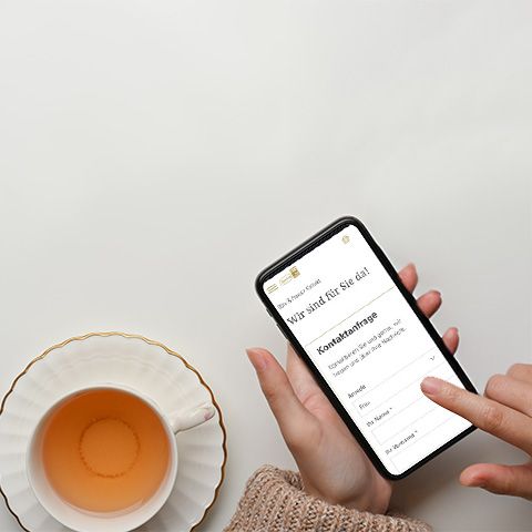 Person nutzt Smartphone, um ein Online-Kontaktformular auszufüllen, neben einer Tasse Tee.