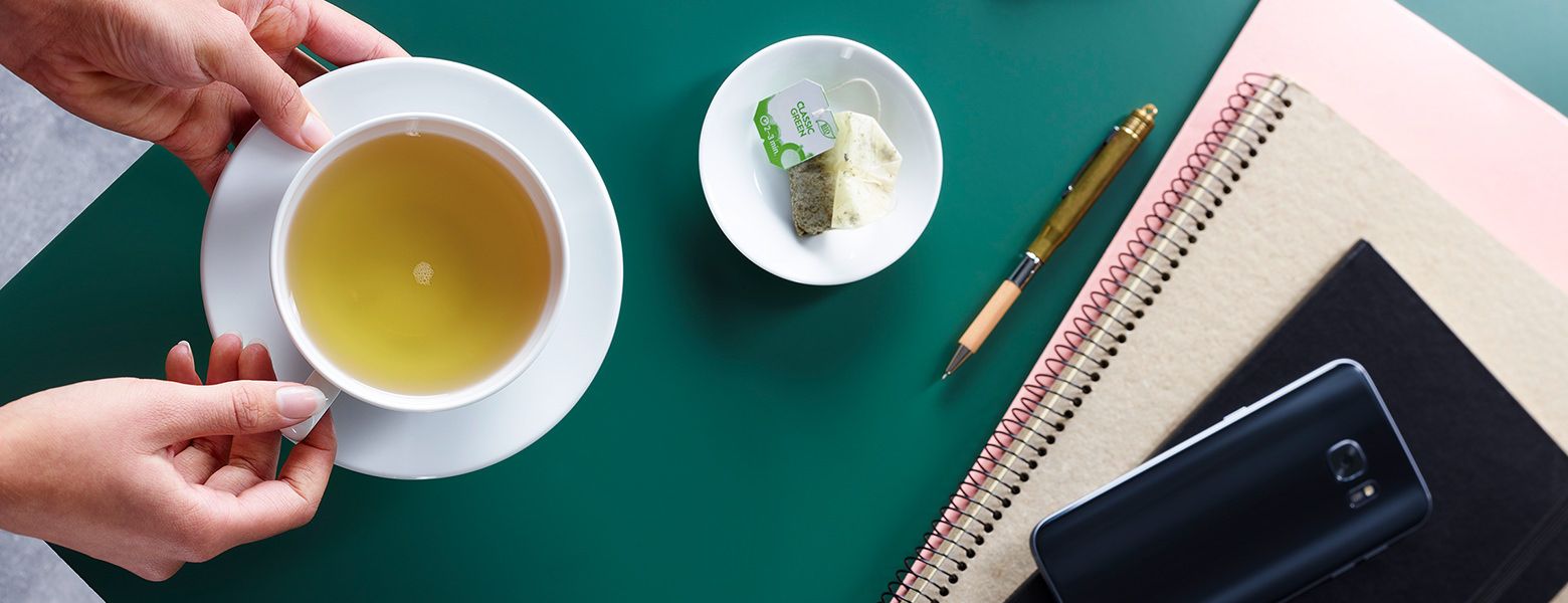 Tasse grüner Tee auf Schreibtisch mit Notizbuch, Stift und Teebeutel.