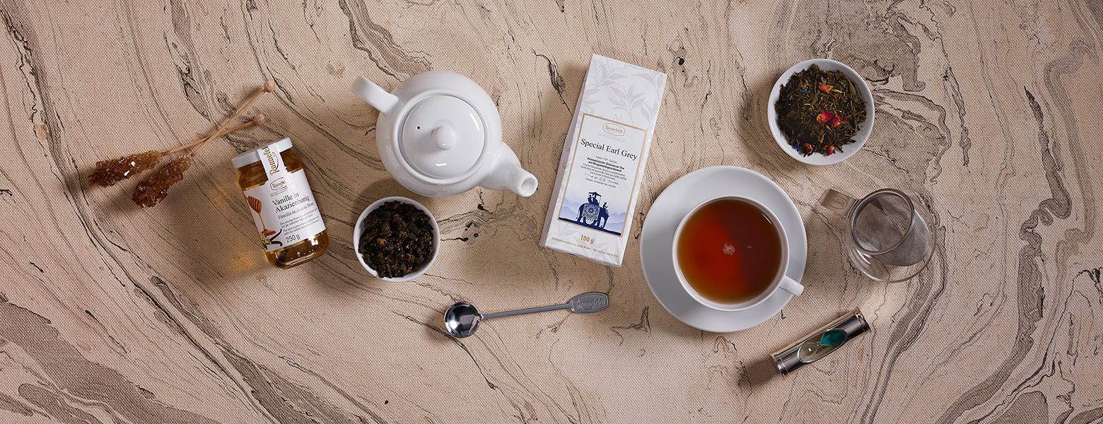 Flach gelegtes Teeset mit Tasse Earl Grey Tee, Teekanne, losem Tee, Teeverpackung, Honig, Kandiszucker, Sanduhr, Teelöffel und Glasfilter auf marmorierter Oberfläche.