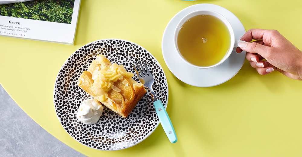 Tasse grüner Tee und ein Stück Apfelkuchen mit Sahne auf modernem gelbem Tisch.