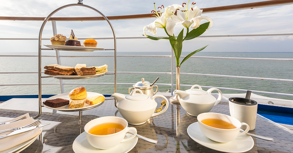 Afternoon-Tea-Setup auf dem Schiffsdeck mit Meerblick, serviert mit Tee, Gebäck und einer Etagere mit süßen und herzhaften Snacks.