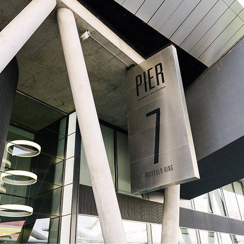 Moderne Gebäudeansicht mit großem Schild „PIER 7“ an schrägen Betonstützen, Standortangabe „Rotfeder-Ring“.