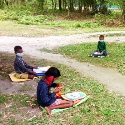 Drei Kinder mit Masken sitzen mit Abstand auf dem Boden im Freien und lernen aus Heften, umgeben von Natur