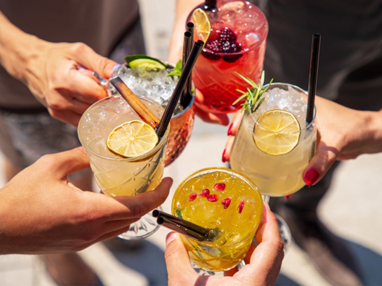 Menschen stoßen mit bunten Sommerdrinks an – Gläser mit Eis, Limetten, Beeren, Granatapfelkernen und Kräutern