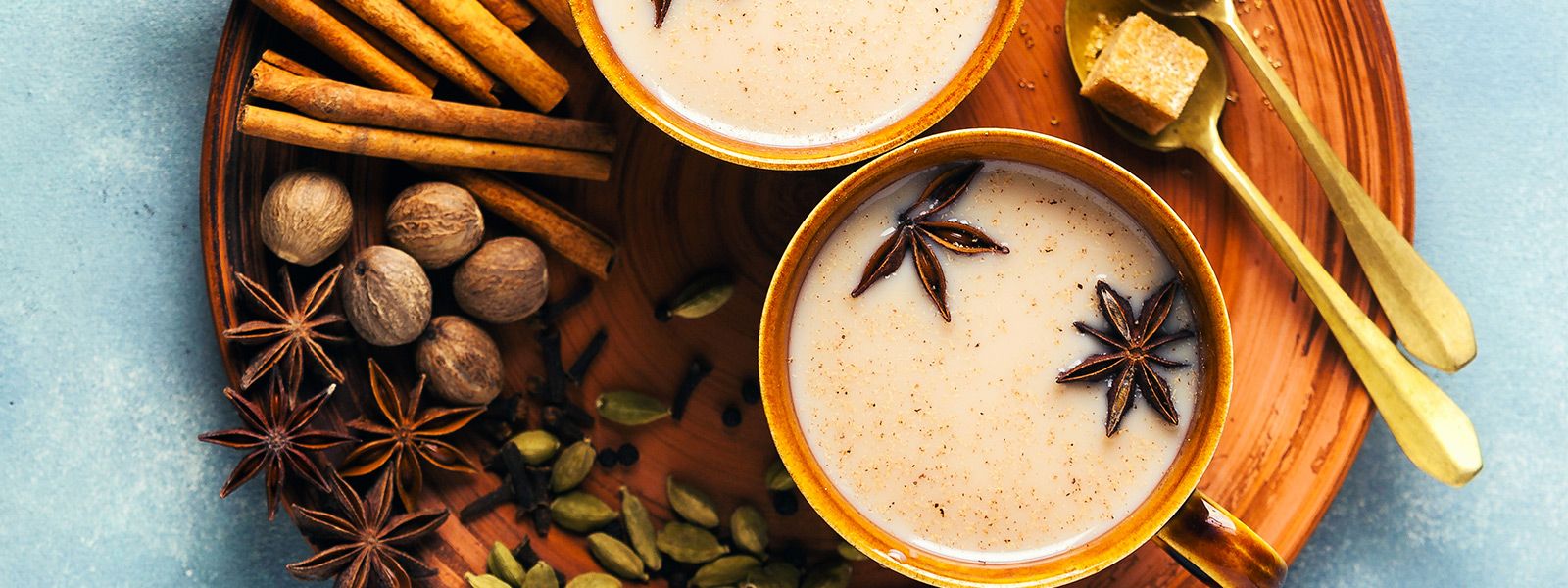 Zwei Tassen Chai-Tee mit Sternanis, umgeben von Gewürzen wie Zimt, Kardamom und Muskatnuss auf einem Holztablett