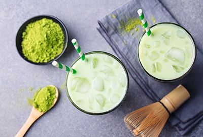 Zwei Gläser Iced Matcha Latte mit Eiswürfeln, Matchapulver und Bambusbesen auf grauem Untergrund