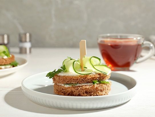 Kleines Sandwich mit Gurken und Kräutern auf einem weißen Teller, daneben eine Tasse Tee