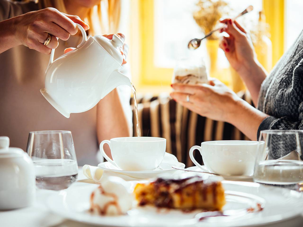 Zwei Frauen genießen gemeinsam eine Teepause, während Tee in weiße Tassen gegossen wird und Kuchen auf dem Tisch steht
