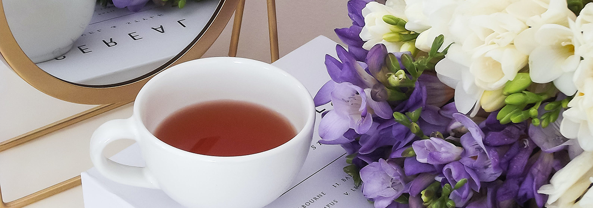Weiße Teetasse mit dem Tee neben einem Spiegel und einem Strauß weiß-lila Blumen