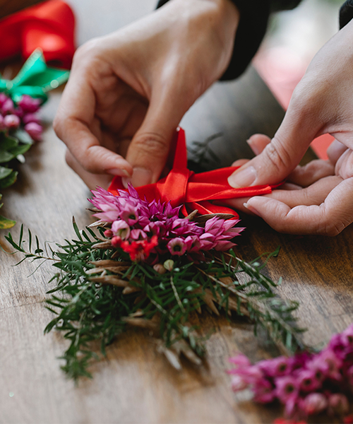 Hände binden eine rote Schleife an ein kleines Gesteck aus rosa Blüten und Tannenzweigen auf einem Holztisch