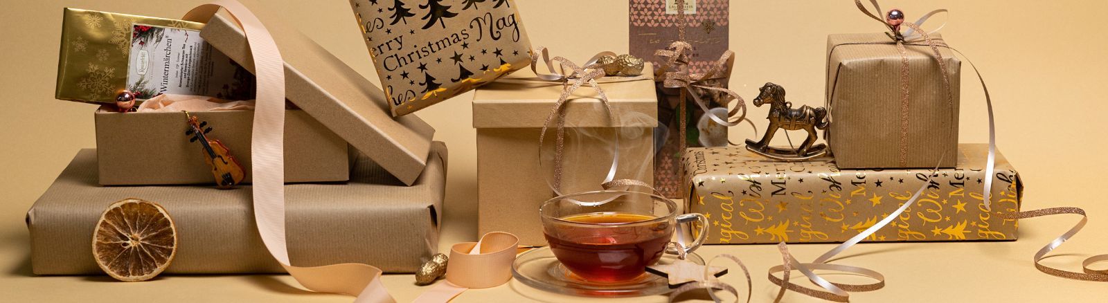 Ronnefeldt Weihnachtsmomente Weihnachtlich verpackte Geschenke, Schleifen und Deko-Elemente arrangiert um eine dampfende Tasse Tee auf beige-goldenem Hintergrund.