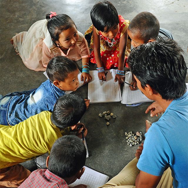 Group of young children sitting in a circle on the floor with a teacher, learning and playing with notebooks and small stones.