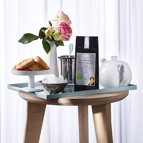 Ein stilvoll gedeckter Holztisch mit einer weißen Teekanne, einer schwarzen Tee-Packung mit der Aufschrift 'Colombian White Imperial', einer Schale mit Zuckerwürfeln, einem Besteckhalter mit Löffel, einer Vase mit Rosen und einem Teller mit Keksen.