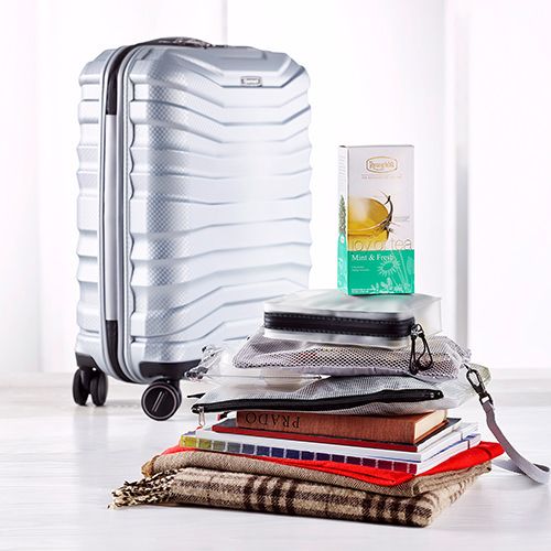 Silberner Reisekoffer im Hintergrund, davor ein Stapel Bücher mit einer Packung Ronnefeldt Joy of Tea Mint & Fresh, sowie Reiseutensilien wie eine Tasche und ein Schal.