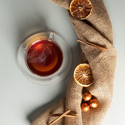 Glastasse mit schwarzem Tee auf einer Glas-Untertasse, dekoriert mit getrockneten Orangenscheiben, Zimtstangen, goldenen Kugeln und einem Jutestoff auf hellem Hintergrund.