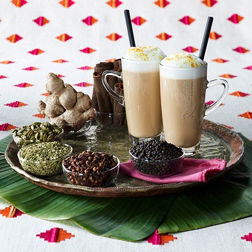 Zwei Gläser Chai-Tee mit Milchschaum und Strohhalmen auf einem verzierten Tablett mit Gewürzen wie Ingwer, Zimtstangen, Kardamom, Nelken und Pfefferkörnern, auf grünen Blättern vor einem farbenfrohen Stoffhintergrund.