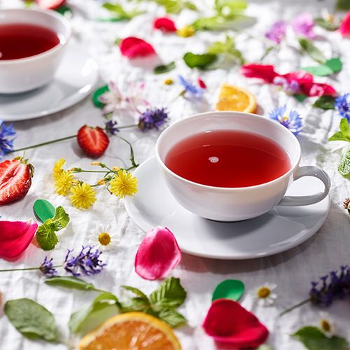 Weiße Teetasse mit rotem Früchtetee auf einer weißen Tischdecke, dekoriert mit bunten Blüten, Erdbeeren, Orangenscheiben und einer zweiten Teetasse im Hintergrund.