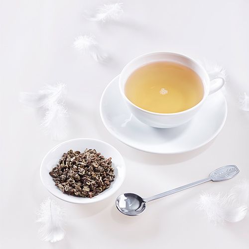 Weiße Teetasse mit weißem Tee auf hellem Hintergrund, daneben eine kleine Schale mit losem Tee, ein silberner Teelöffel und weiße Federn.