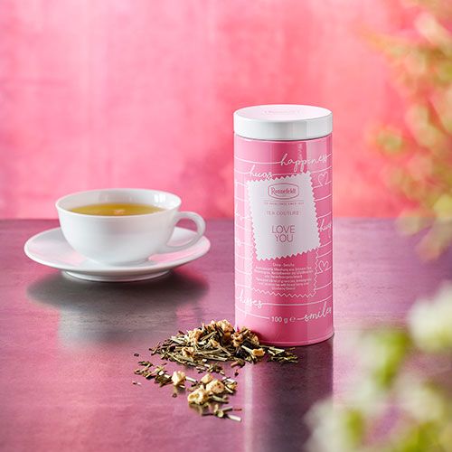 Eine rosa Teedose mit der Aufschrift 'I LOVE YOU' steht auf einem Tisch, daneben eine weiße Teetasse mit heißem Tee und lose Teeblätter. Der Hintergrund ist pink gehalten.