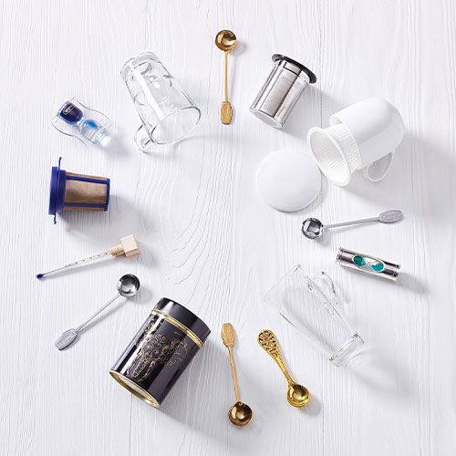 Verschiedene Ronnefeldt Tee-Accessoires wie Teesiebe, Messlöffel, Gläser und Teedosen auf weißem Untergrund kreisförmig angeordnet.